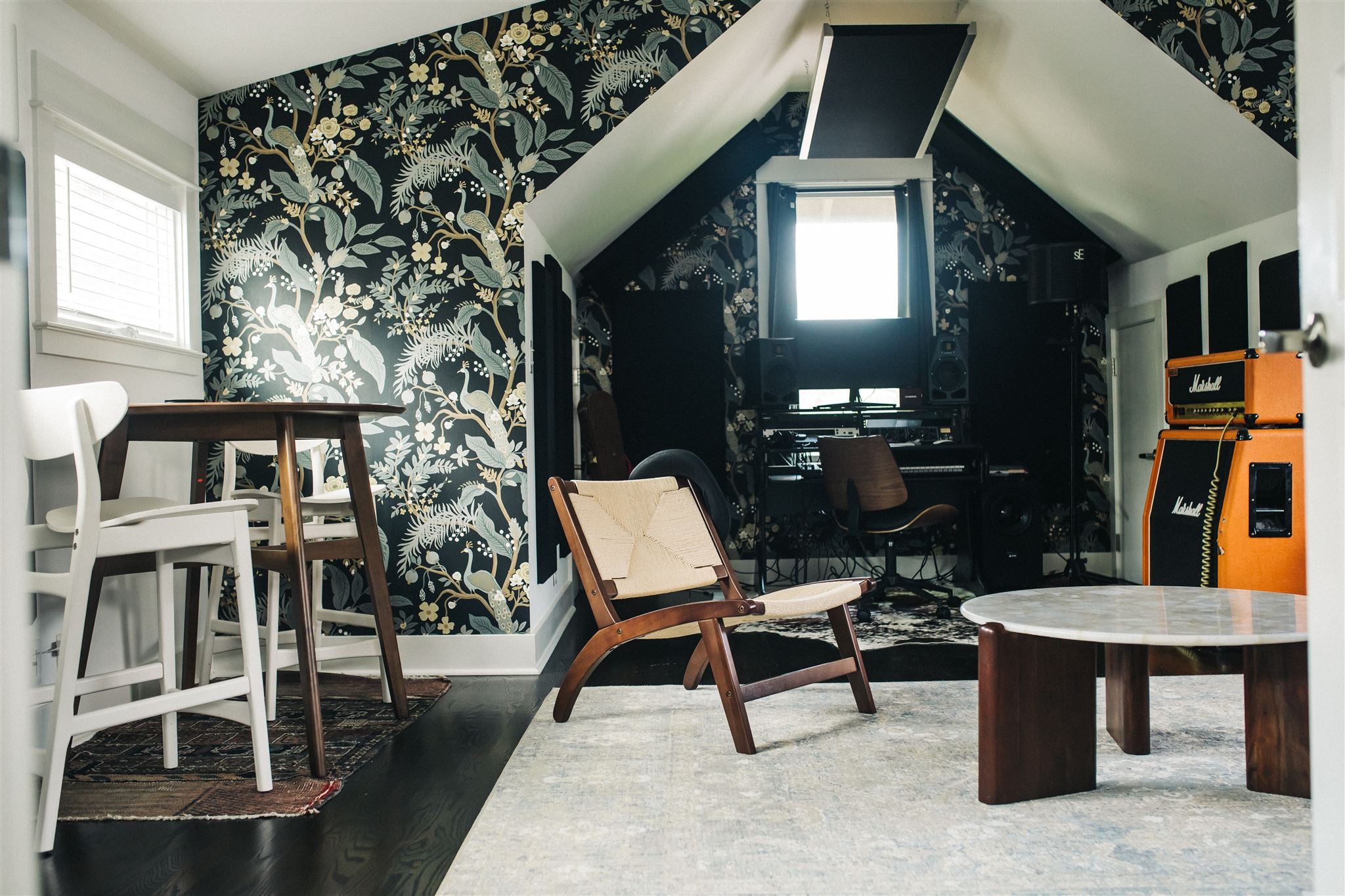 Interior of a cozy attic recording studio with dark botanical-print wallpaper, wooden chairs, a round marble coffee table, and a Marshall amp stack visible in the background near recording equipment
