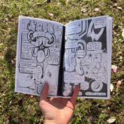 A hand holding open an illustrated lyric zine outdoors over grass, showing black and white hand-drawn comic-style artwork with "Track #3" in graffiti lettering on the left page and lyric snippets with cartoon characters on the right