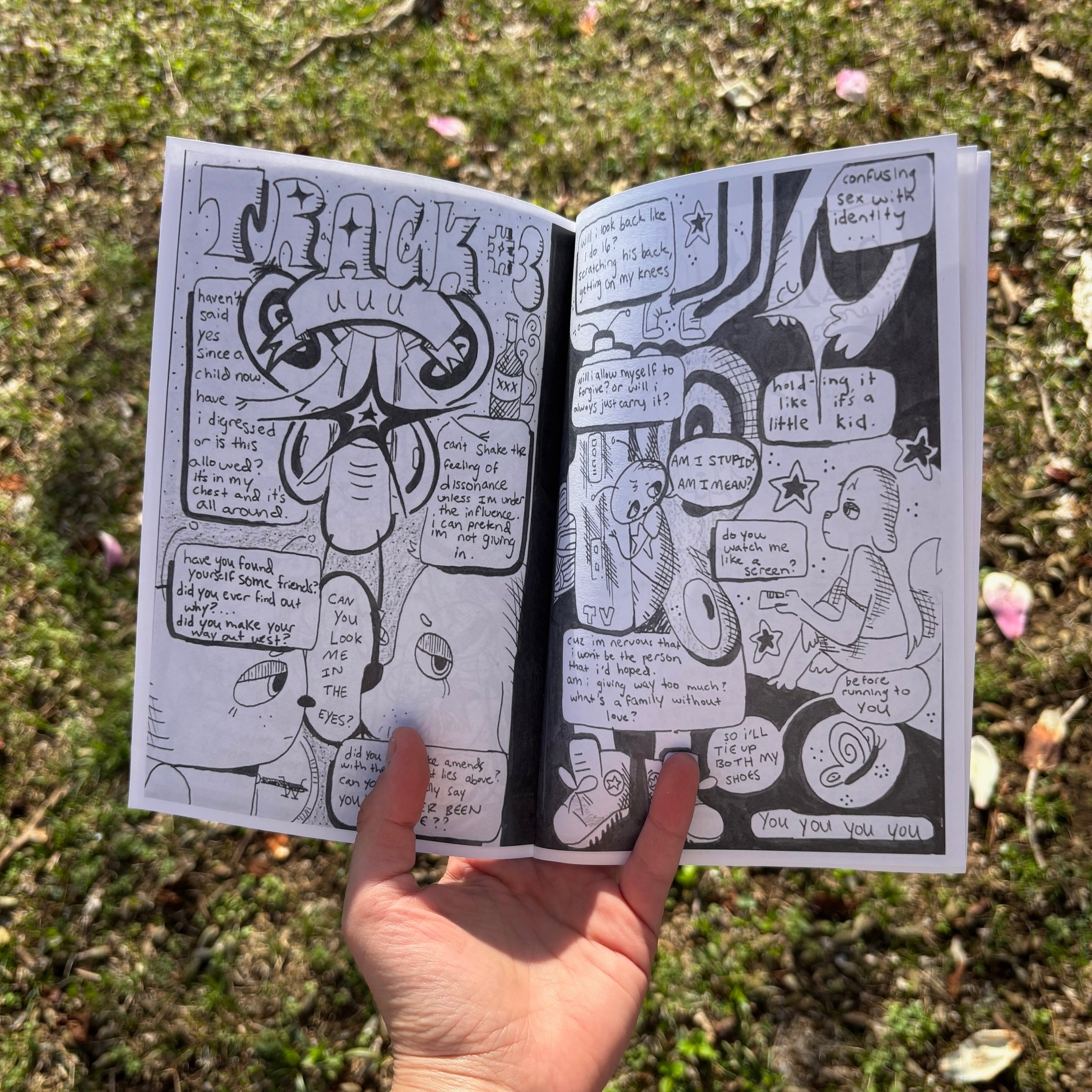 A hand holding open an illustrated lyric zine outdoors over grass, showing black and white hand-drawn comic-style artwork with "Track #3" in graffiti lettering on the left page and lyric snippets with cartoon characters on the right