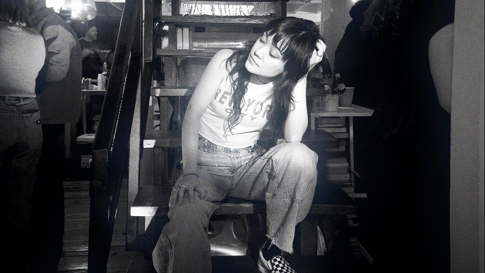 Apple Music thumbnail for Ok Cowgirl's "I Want You" — black and white photo of Ok Cowgirl sitting on stairs in a bar or venue setting, wearing a graphic tank top, jeans, and checkered sneakers, with other patrons visible in the background