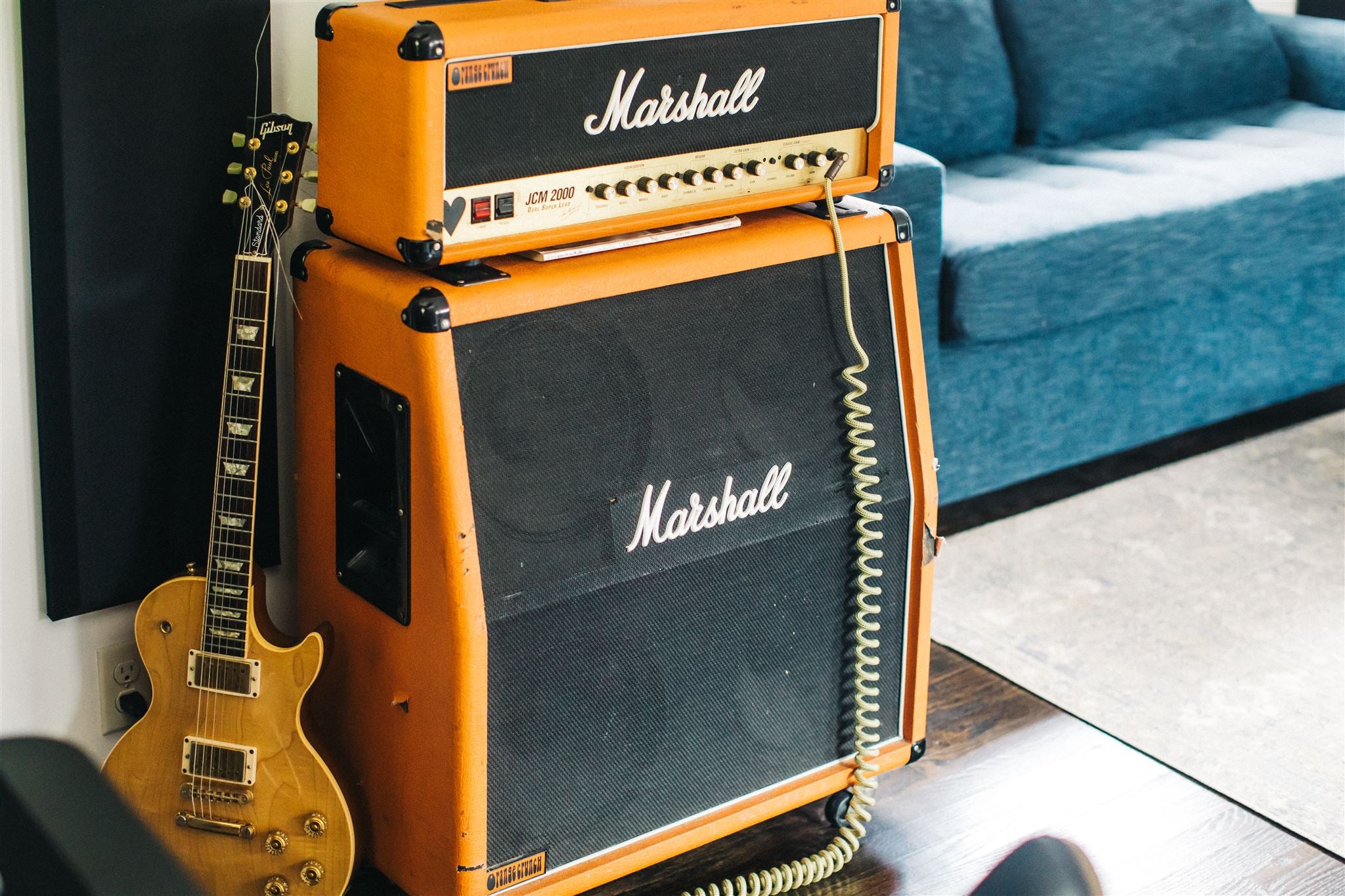 A Marshall JCM 2000 guitar amplifier stack in orange finish with a Gibson Les Paul electric guitar leaning against it, in a home studio or living room setting with a couch in the background