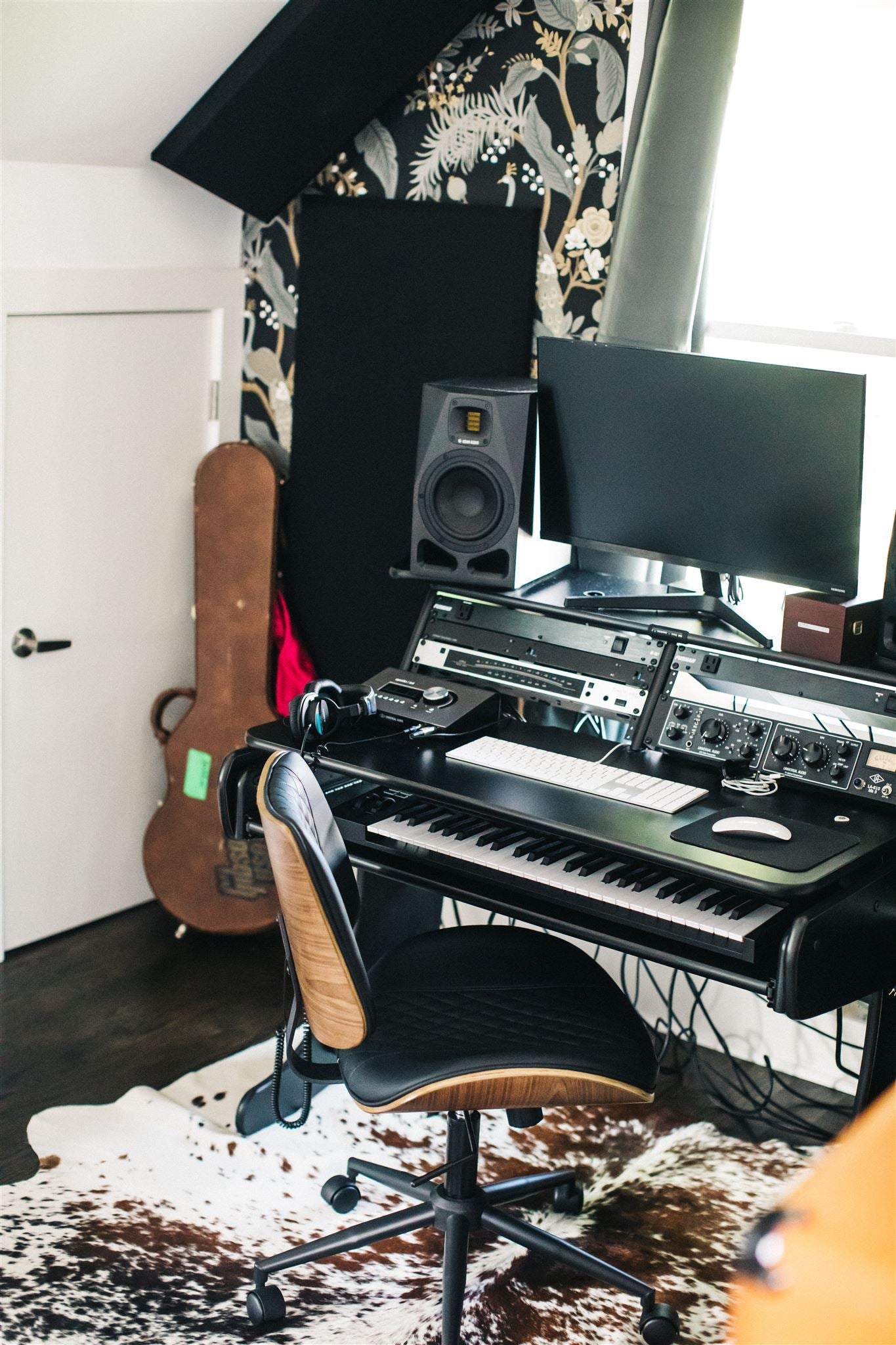 A home recording studio corner with a keyboard workstation, studio monitor speakers, computer monitor, audio interface, and acoustic foam panels, with dark botanical wallpaper and an acoustic guitar visible in the background